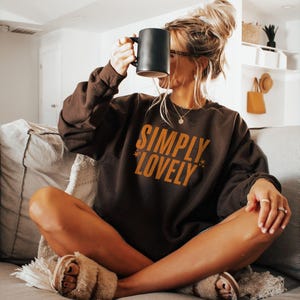 Pu&ograve; includere: Una felpa marrone con la scritta "SIMPLY LOVELY" in arancione. La persona tiene in mano una tazza nera. La persona &egrave; seduta su un divano con una coperta e cuscini di colore chiaro. La persona indossa pantofole soffici.