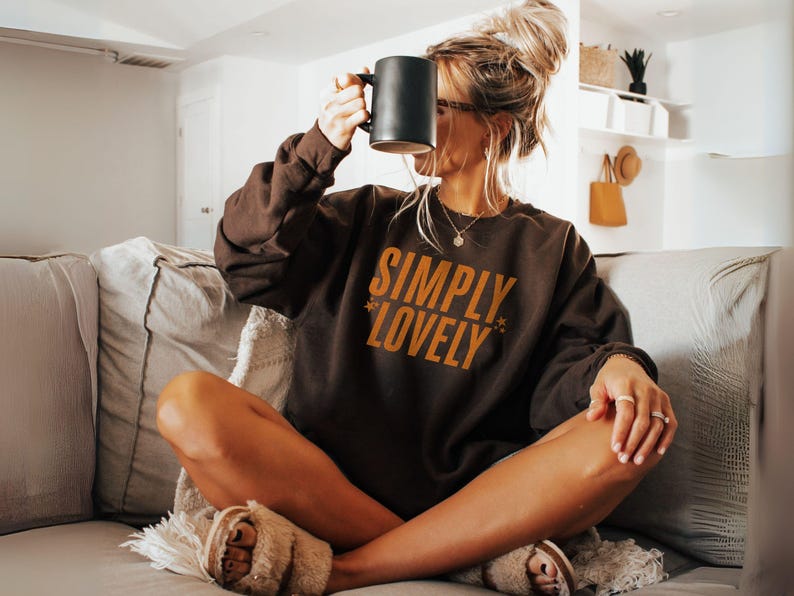 Pu&ograve; includere: Una felpa marrone con la scritta "SIMPLY LOVELY" in arancione. La persona tiene in mano una tazza nera. La persona &egrave; seduta su un divano con una coperta e cuscini di colore chiaro. La persona indossa pantofole soffici.