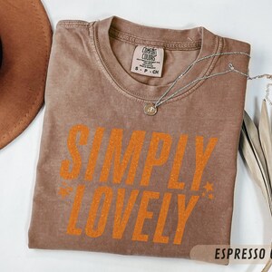 May include: A brown t-shirt in the colour 'Espresso' with the words "SIMPLY LOVELY" in orange. A silver necklace with a pendant lies on the shirt. A brown hat and dried plant are in the background.