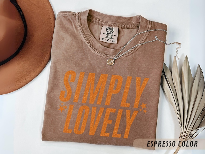 May include: A brown t-shirt in the colour 'Espresso' with the words "SIMPLY LOVELY" in orange. A silver necklace with a pendant lies on the shirt. A brown hat and dried plant are in the background.