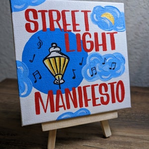 May include: A hand-painted canvas with the words "Street Light Manifesto" in red lettering. The background is blue with white clouds and musical notes. A yellow streetlight is in the center of the canvas.