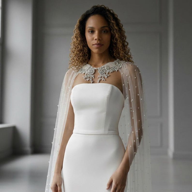 May include: A white strapless gown featuring a sheer tulle capelet adorned with scattered white pearls. The capelet includes a prominent, intricate collar embellished with clear crystals and silver-toned beads, draping over the shoulders.