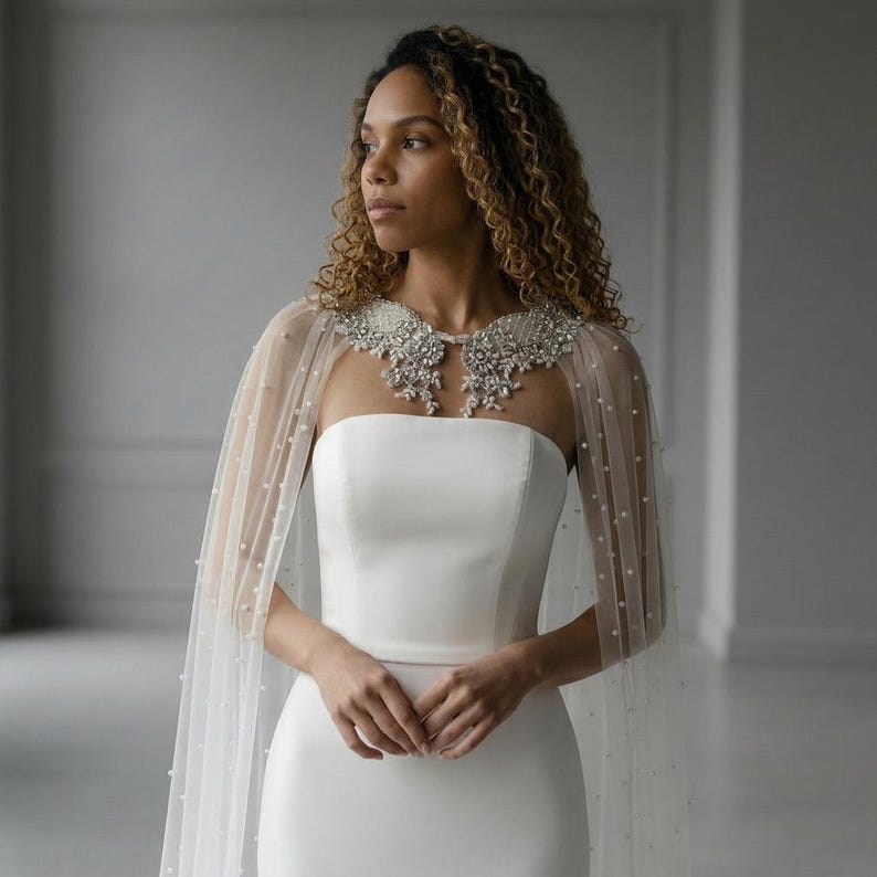 May include: A strapless white gown paired with a sheer, floor-length cape featuring scattered white pearls. The cape includes an intricate, silver-toned beaded collar that rests across the shoulders and neckline, adding texture to the smooth fabric.