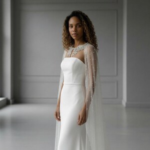 May include: A floor-length white strapless gown featuring a fitted bodice and a long, sheer tulle cape overlay. The cape is adorned with scattered small white pearls and features an intricate, beaded lace collar detail around the neckline.