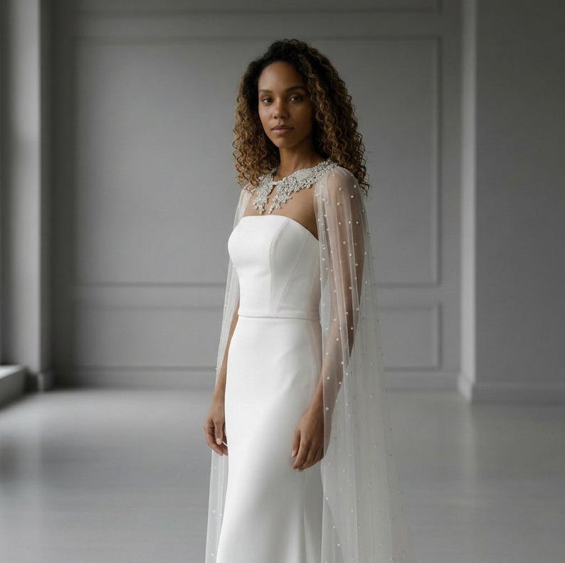 May include: A floor-length white strapless gown featuring a fitted bodice and a long, sheer tulle cape overlay. The cape is adorned with scattered small white pearls and features an intricate, beaded lace collar detail around the neckline.