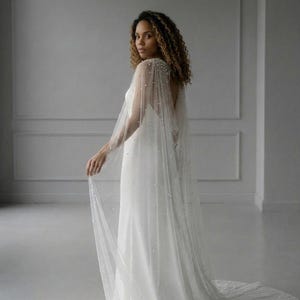 May include: A floor-length white bridal cape made of sheer tulle fabric, featuring scattered white pearl embellishments throughout the material. The cape attaches at the shoulders and drapes down the back, creating a long, flowing silhouette.