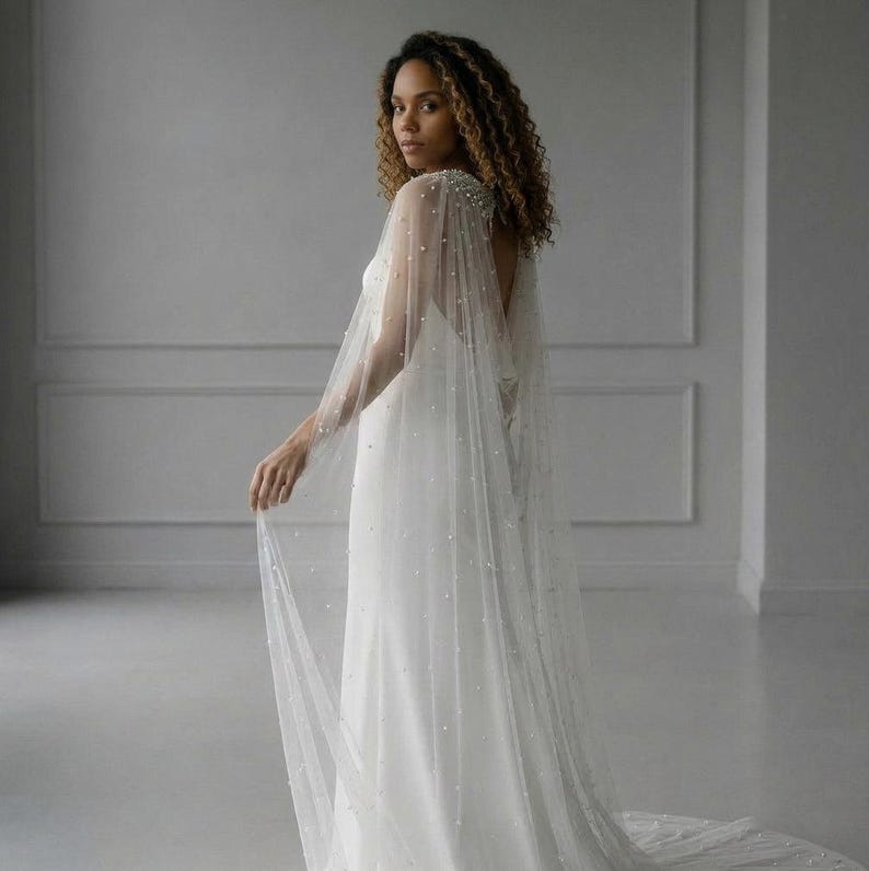 May include: A floor-length white bridal cape made of sheer tulle fabric, featuring scattered white pearl embellishments throughout the material. The cape attaches at the shoulders and drapes down the back, creating a long, flowing silhouette.