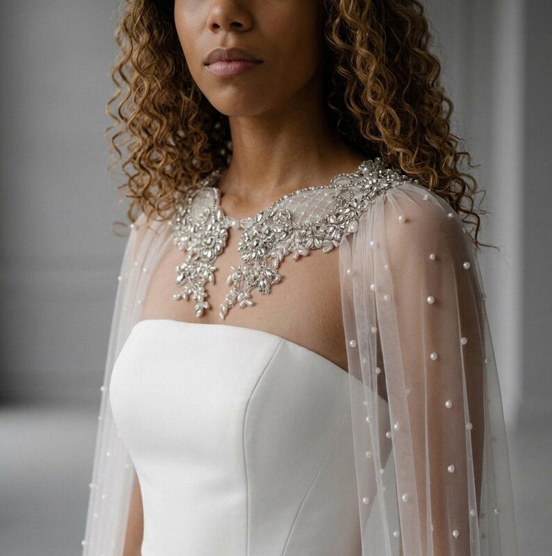 May include: A sheer white tulle capelet featuring scattered white pearls and an intricate collar embellished with silver-toned crystals and beads. The capelet is worn over a strapless white garment, highlighting the detailed embroidery and beadwork.