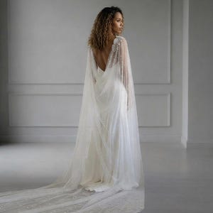May include: A floor-length white gown featuring a deep open back and a sheer, flowing cape overlay. The cape is adorned with scattered white pearls and intricate beaded detailing at the shoulders, extending into a long, sweeping train.