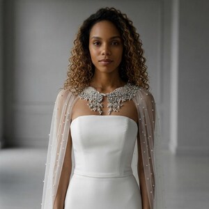 May include: A white strapless gown featuring a sheer tulle capelet adorned with scattered white pearls. The capelet is accented by an intricate, silver-toned collar embellished with clear crystals and beads that drape over the shoulders.