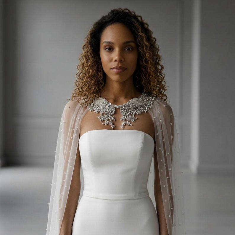 May include: A white strapless gown featuring a sheer tulle capelet adorned with scattered white pearls. The capelet is accented by an intricate, silver-toned collar embellished with clear crystals and beads that drape over the shoulders.