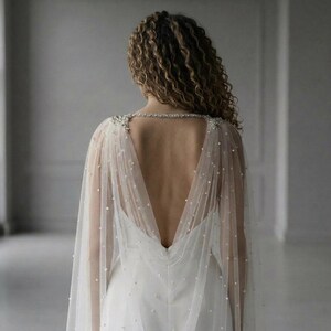 May include: A sheer, floor-length bridal capelet in ivory tulle, featuring scattered white pearls throughout the fabric. The capelet attaches at the shoulders with intricate silver-toned crystal embellishments and drapes over the back.