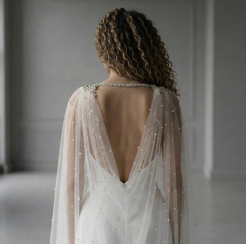 May include: A sheer, floor-length bridal capelet in ivory tulle, featuring scattered white pearls throughout the fabric. The capelet attaches at the shoulders with intricate silver-toned crystal embellishments and drapes over the back.