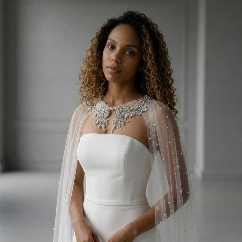 May include: A white strapless gown paired with a sheer tulle capelet featuring scattered white pearls. The capelet includes a prominent, intricate collar embellished with silver-toned embroidery, clear crystals, and hanging pearl accents.