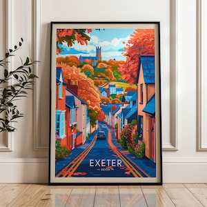 Op de afbeelding: Een ingelijste kunstafdruk van Exeter, Devon, met een kleurrijke straatscène. Het kunstwerk toont een weg met gebouwen en een kerk op de achtergrond. De afdruk heeft een zwarte lijst en wordt op een houten vloer gepresenteerd.
