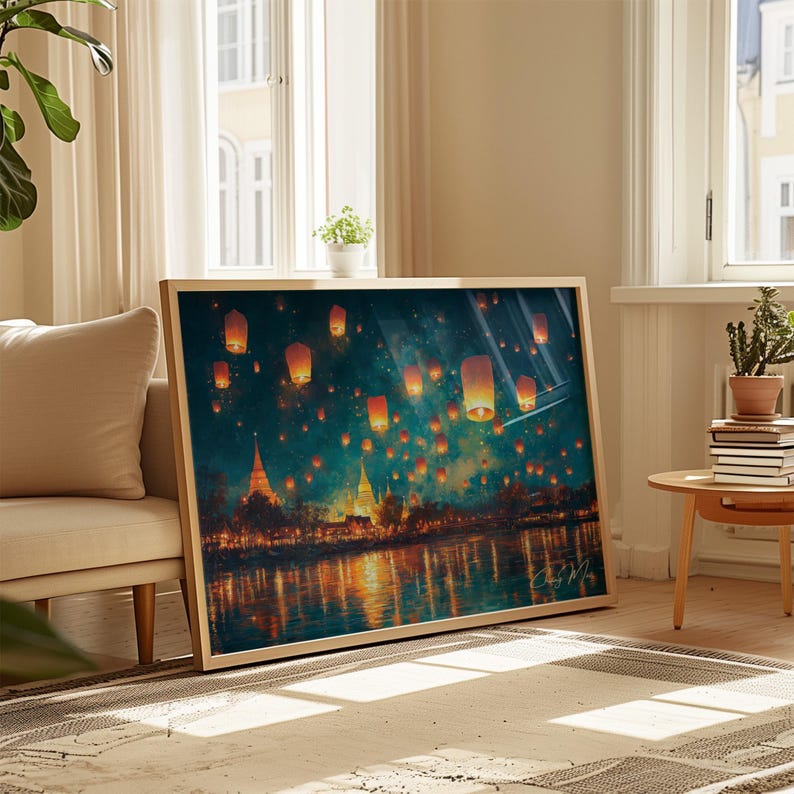 May include: A framed art print showing a night scene with floating lanterns over water. The artwork features orange and yellow lanterns against a dark blue and teal sky, with reflections in the water. The frame is a light wood colour.