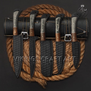 May include: Set of six black kitchen knives with textured blades and wooden handles, displayed in a black leather roll on a brown rope background. The knives are arranged in a row, with the handles facing the viewer. The roll is closed, and the knives are secured with a buckle.
