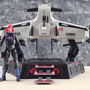 May include: A white and black Cobra aircraft with the word "COBRA" printed on the side, and a red and black base. A figure in black and blue armor and a red helmet stands next to the aircraft.