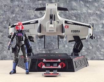 May include: A white and black Cobra aircraft with the word "COBRA" printed on the side, and a red and black base. A figure in black and blue armor and a red helmet stands next to the aircraft.