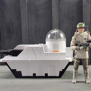 May include: A white toy tank with black details and a clear dome, next to a figure in a white and tan uniform, holding a rifle. The tank has a rectangular shape with a black tread base and a black gun barrel.