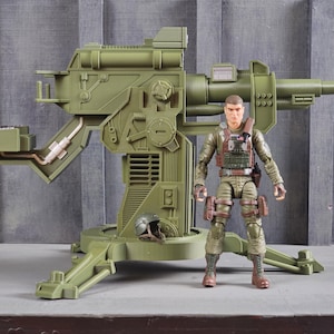 May include: An olive green toy artillery cannon with a detailed design, standing next to a military action figure. The figure is wearing a green uniform and brown boots. A helmet rests on the base of the cannon.