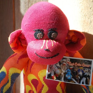 Puede incluir: Un mono de calcetín rojo con llamas amarillas y naranjas en su cuerpo. El mono sostiene una tarjeta que dice "Sock Critter Corner" y tiene una imagen de varios monos de calcetín.