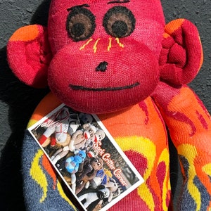 Puede incluir: Un mono de calcetín rojo con un patrón de llamas amarillas y naranjas. El mono sostiene una tarjeta que dice "Sock Critter Corner".