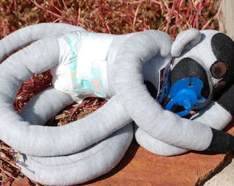May include: A gray and black sock monkey toy wearing a diaper and holding a blue pacifier. The monkey is lying on its side with its head resting on its arm.