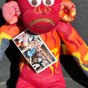 Puede incluir: Un mono de calcetín rojo con un patrón de llamas, que lleva una etiqueta blanca que dice "Sock Critter Corner".