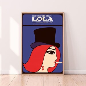 May include: A vintage film poster for the movie "Lola" starring Anouk Aimée. The poster features a stylised illustration of a woman with red hair wearing a black top hat and smoking a cigarette. The background is a solid blue colour.