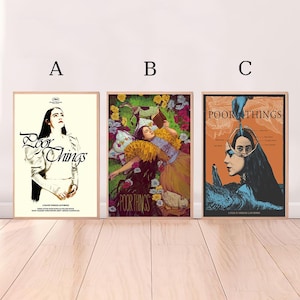 May include: Three framed movie posters for "Poor Things" are displayed. The posters feature different artistic styles, with the film's title prominently displayed. The posters are in wooden frames and are set against a white wall.