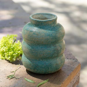 May include: A small, bulbous ceramic vase with a textured, teal-colored glaze featuring subtle brown speckles. The vase has a distinct three-tiered, rounded shape with a narrow opening at the top and a smooth, slightly uneven surface finish.