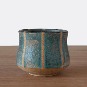 May include: A handcrafted ceramic bowl with a teal glaze and vertical gold stripes. The bowl has a rounded shape with a textured surface and a natural, unglazed base. The interior is dark, and the bowl sits on a wooden surface.
