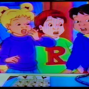 May include: A still from an animated cartoon featuring four children with surprised expressions. One child wears a green shirt with a red "R", another a blue sweater, and the others wear red and yellow outfits. A telephone and a control panel are visible.