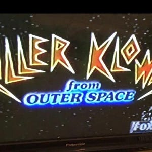 May include: A television screen displaying the title "Killer Klowns from Outer Space" in a stylized font with orange, yellow, and white letters against a black background with white dots. The text "from Outer Space" is in blue.