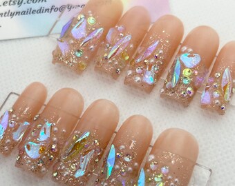 May include: Set of ten nude-colored press-on nails decorated with iridescent glitter, iridescent rhinestones, and small white pearls. The nails are arranged on a clear plastic display card.