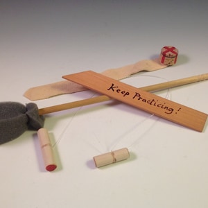 May include: A set of wooden juggling equipment with a gray foam ball, a wooden stick, and two wooden cylinders. The equipment is on a white surface with a wooden sign that says "Keep Practicing!"