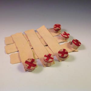 May include: Five cork bottle stoppers with a red cross design on top, arranged on a beige fabric surface.