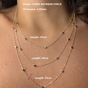 May include: Three gold necklaces with square-shaped beads. The necklaces are layered on a person's neck and are available in three lengths: 45cm, 50cm, and 55cm. The necklaces are 3mm thick. Model: CUBED BETWEEN FORCE.