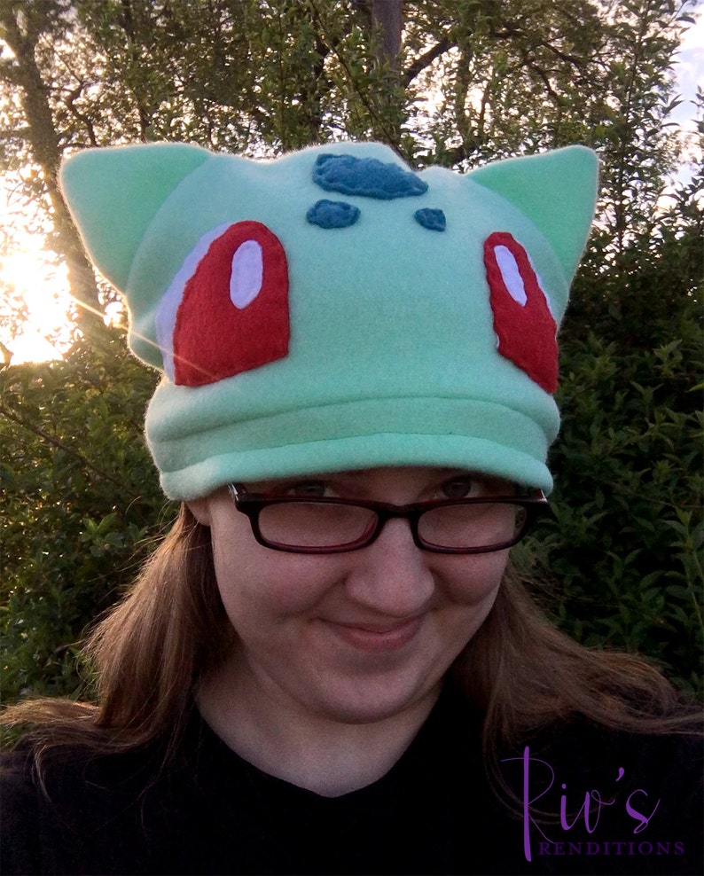 Pu&ograve; includere: Un cappello in pile verde chiaro con grandi orecchie a punta e accenti rossi e bianchi, simile a un personaggio di Bulbasaur. Il cappello ha dettagli blu sulla fronte.