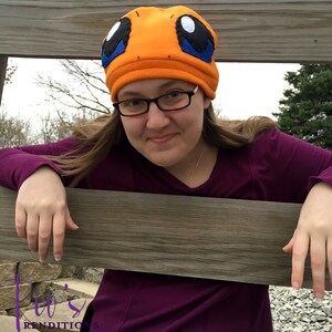 May include: An orange fleece hat with a cartoon-style face, featuring large black, blue, and white eyes. The hat is worn by a person wearing glasses and a purple long-sleeved shirt. The hat is a handmade item.