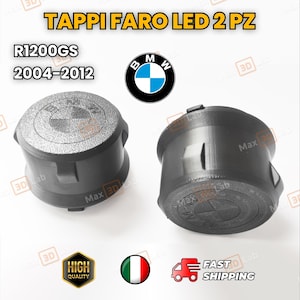 Può includere: Due tappi neri per fari a LED con la scritta "TAPPI FARO LED 2 PZ" e "R1200GS 2004-2012". I tappi hanno il logo BMW e sono etichettati "Max 3D Lab". L'immagine include anche un badge "High Quality".