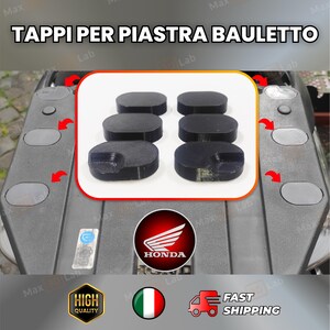 Può includere: Pezzi di plastica neri di forma ovale, possibilmente per un veicolo, sono esposti in una scatola bordata di bianco. Il testo "TAPPI PER PIASTRA BAULETTO" è in alto. Il logo Honda è visibile sotto, insieme ad altre icone.