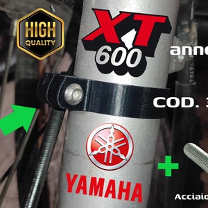 Può includere: Porta cavo nero per una moto Yamaha XT 600. Il supporto è in metallo e ha una vite per fissarlo alla moto. Il testo "COD. 34L-23318-01" è visibile sul supporto. Il testo "VITE INCLUSA" è anche visibile, indicando che è inclusa una vite.