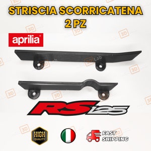 Puede incluir: Dos protectores de cadena de plástico negro para una motocicleta. Los protectores están diseñados para proteger la cadena de la suciedad y los residuos. Los protectores están etiquetados con "STRISCIA SCORRICAΤΕΝΑ 2 PZ" y "aprilia".