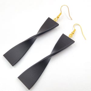 Può includere: Un paio di orecchini pendenti neri attorcigliati con ganci dorati. Gli orecchini hanno un design geometrico semplice e sono su uno sfondo bianco. Un accessorio di moda.