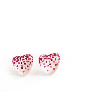 May include: A pair of heart-shaped earrings. Each earring is clear with a pattern of red and pink dots. The earrings are set against a white background.