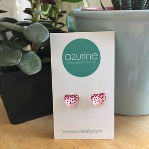 May include: A pair of heart-shaped stud earrings with a white base and red glitter accents. The earrings are displayed on a white card with the brand name "azurine" and the website address www.azurine.etsy.com.