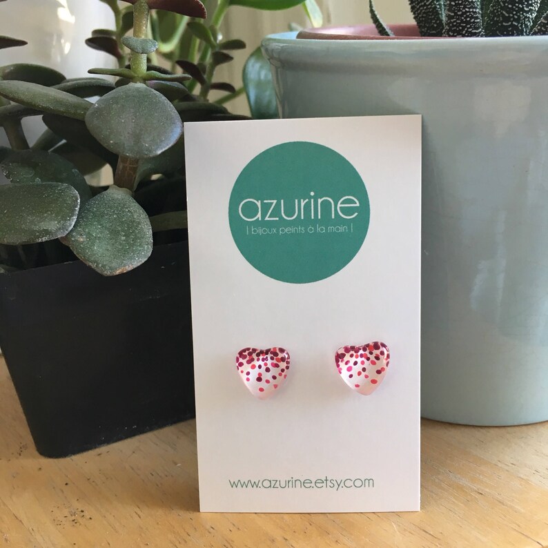 May include: A pair of heart-shaped stud earrings with a white base and red glitter accents. The earrings are displayed on a white card with the brand name "azurine" and the website address www.azurine.etsy.com.