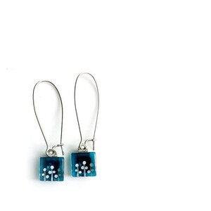 May include: A pair of dangle earrings with silver hooks and blue glass square charms. The charms have a white floral design.
